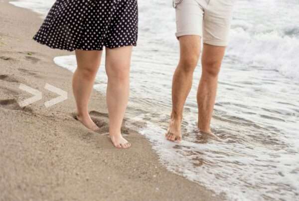 Auf dem Bild sind zwei ältere Menschen zu sehen, die am Strand entlang laufen und dabei barfuß sind.