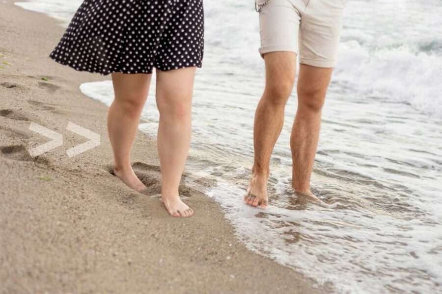 Auf dem Bild sind zwei ältere Menschen zu sehen, die am Strand entlang laufen und dabei barfuß sind.